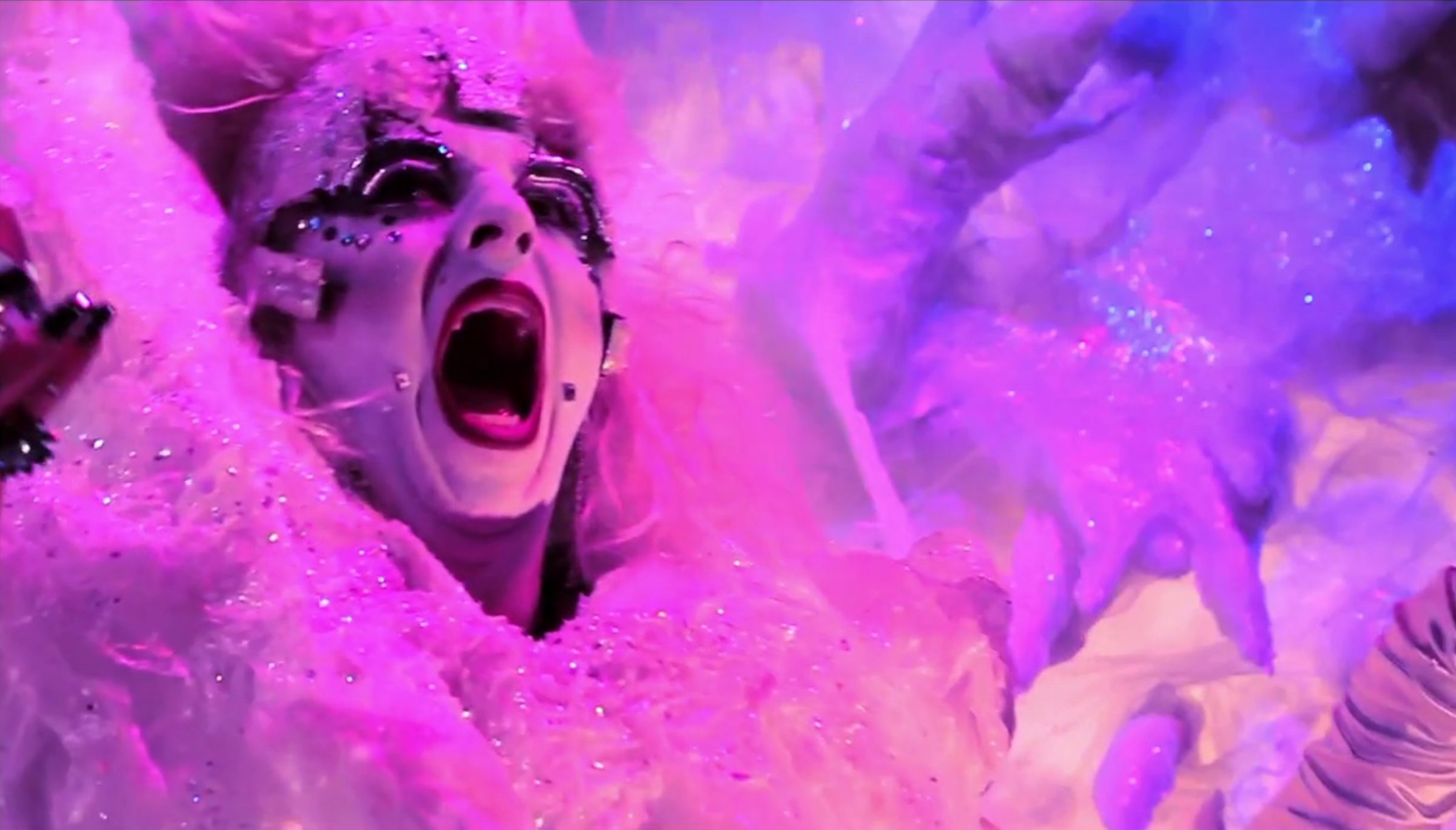 Close-up of a person with dramatic makeup, open mouth, and glitter, surrounded by pink and purple light and fabric.